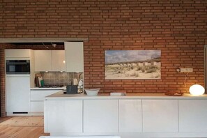 A kitchen with a brick wall, a white countertop, and a painting of a desert landscape. - Holiday home with 2 bedrooms (Retranchement)