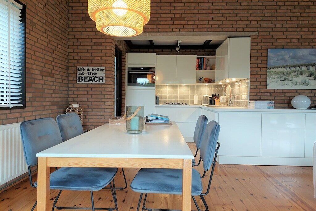 Un coin repas avec une table blanche et des chaises bleues, un mur en briques et une cuisine en arrière-plan.