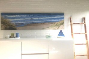 A kitchen with a large beach-themed artwork on the wall, a white countertop, and a bamboo ladder. - Holiday home with 2 bedrooms (Retranchement)