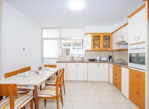 Een keuken met witte kasten, een eettafel met stoelen en een raam met zonwering.