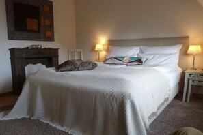 Ein ordentlich bezogenes Bett mit weißer Bettdecke und Kissen, ein kleiner Nachttisch mit Lampe sowie ein Kamin mit Sims.
