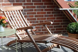 Ein Holzstuhl mit querverlaufenden Leisten, ein runder Metalltisch mit einer weißen Vase und ein Topfpflanzen auf einem Balkon mit Ziegelwand im Hintergrund.