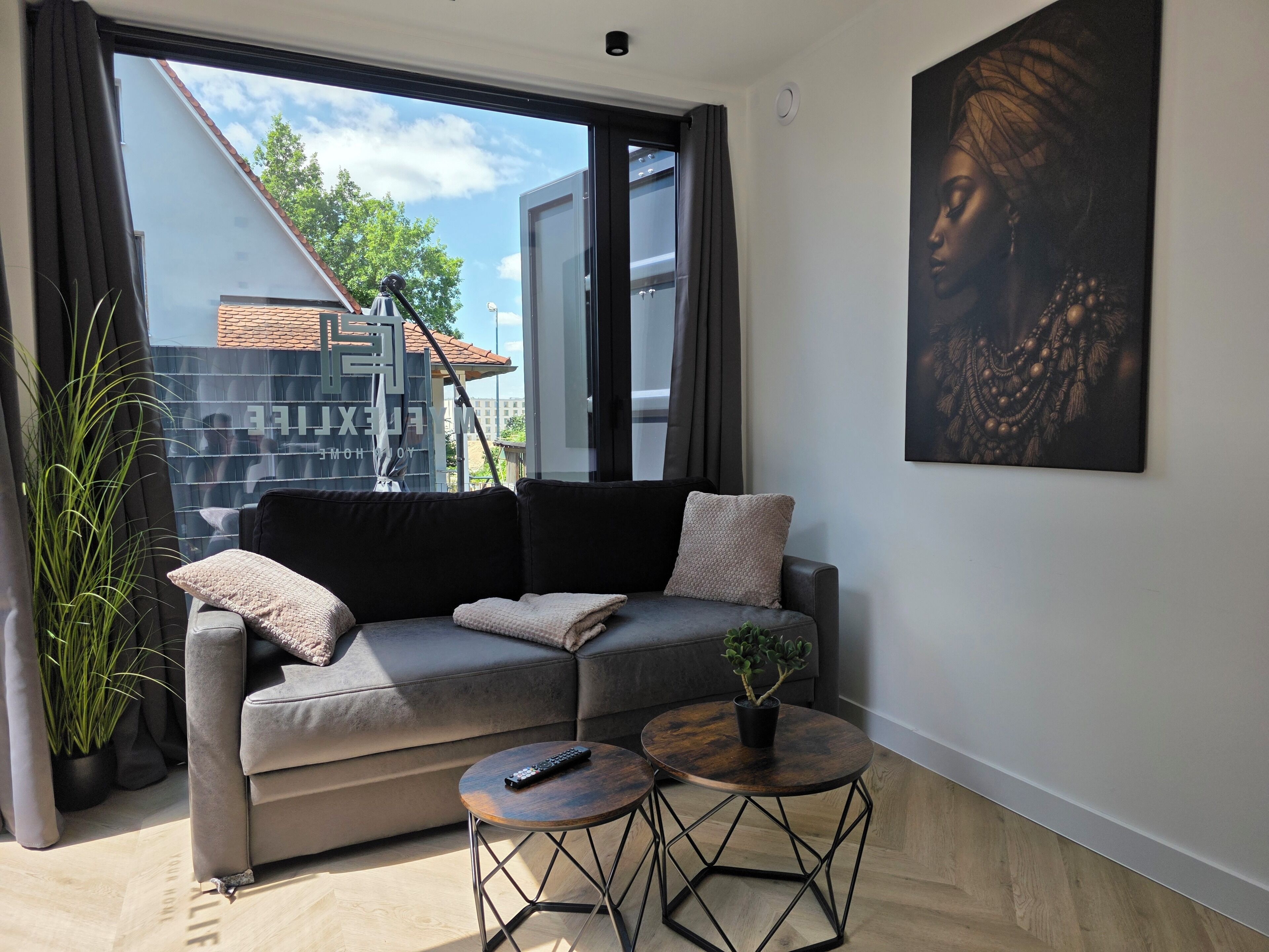 Ein modernes Wohnzimmer mit einem grauen Sofa, zwei runden Tischen, einer Topfpflanze und einem großen Fenster mit Blick auf Gebäude und Bäume.