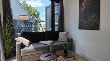 Ein modernes Wohnzimmer mit einem grauen Sofa, zwei runden Tischen, einer Topfpflanze und einem großen Fenster mit Blick auf Gebäude und Bäume.