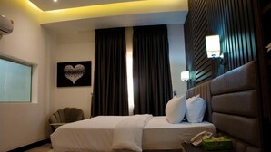 Ein Hotelzimmer mit einem Bett, einem Nachttisch mit Telefon und einer Schachtel, einem Sessel, einer Wandlampe und einem herzförmigen Kunstwerk an der Wand.