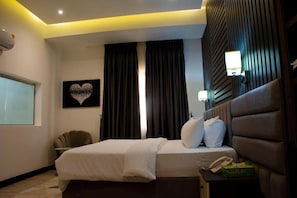 A hotel room with a bed, a bedside table with a phone and a box, a chair, a wall-mounted lamp, and a heart-shaped artwork on the wall. - Aviel Hotel And Suites (Abuja)