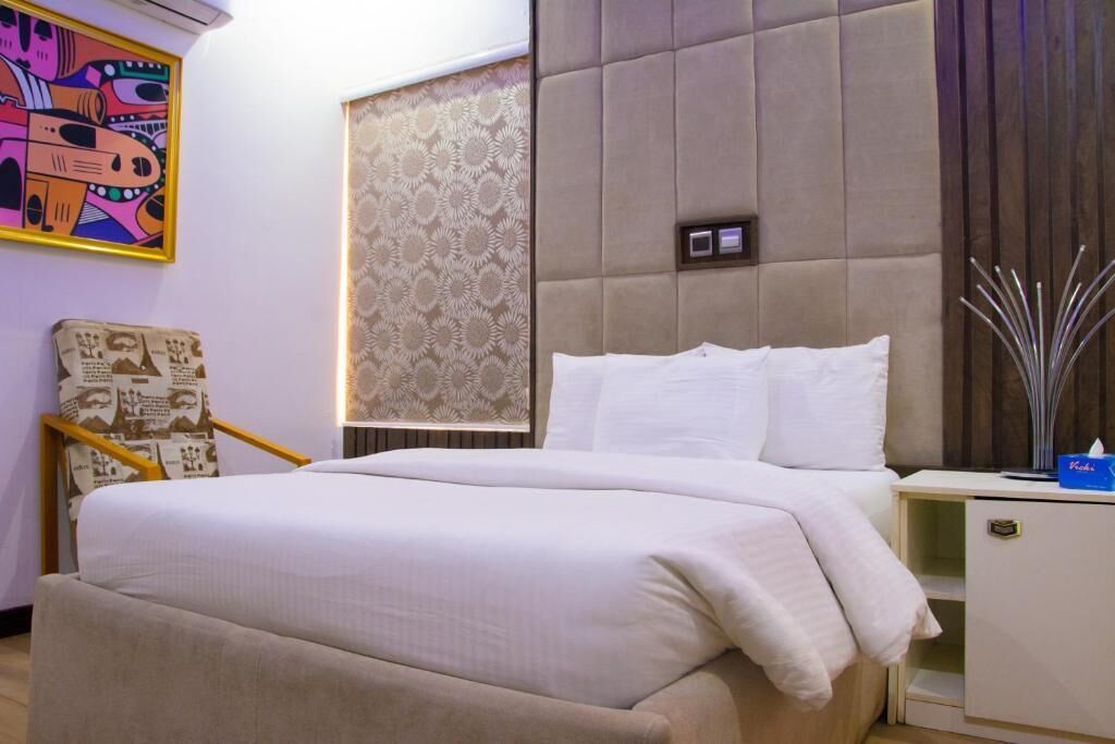 Quarto de hotel com cama, mesa de cabeceira, cadeira e parede com papel de parede estampado e um quadro emoldurado.