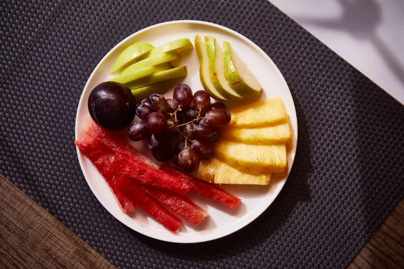 Un piatto con fette di mele, uva, ananas e peperone rosso su un sottopiatti intrecciato.