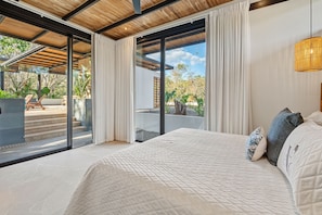 A bedroom with a large bed, white curtains, and a view of a patio with outdoor furniture and plants.