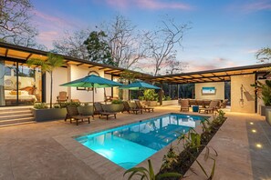 A modern outdoor pool area with lounge chairs and umbrellas, surrounded by a landscaped garden and a building with large windows.