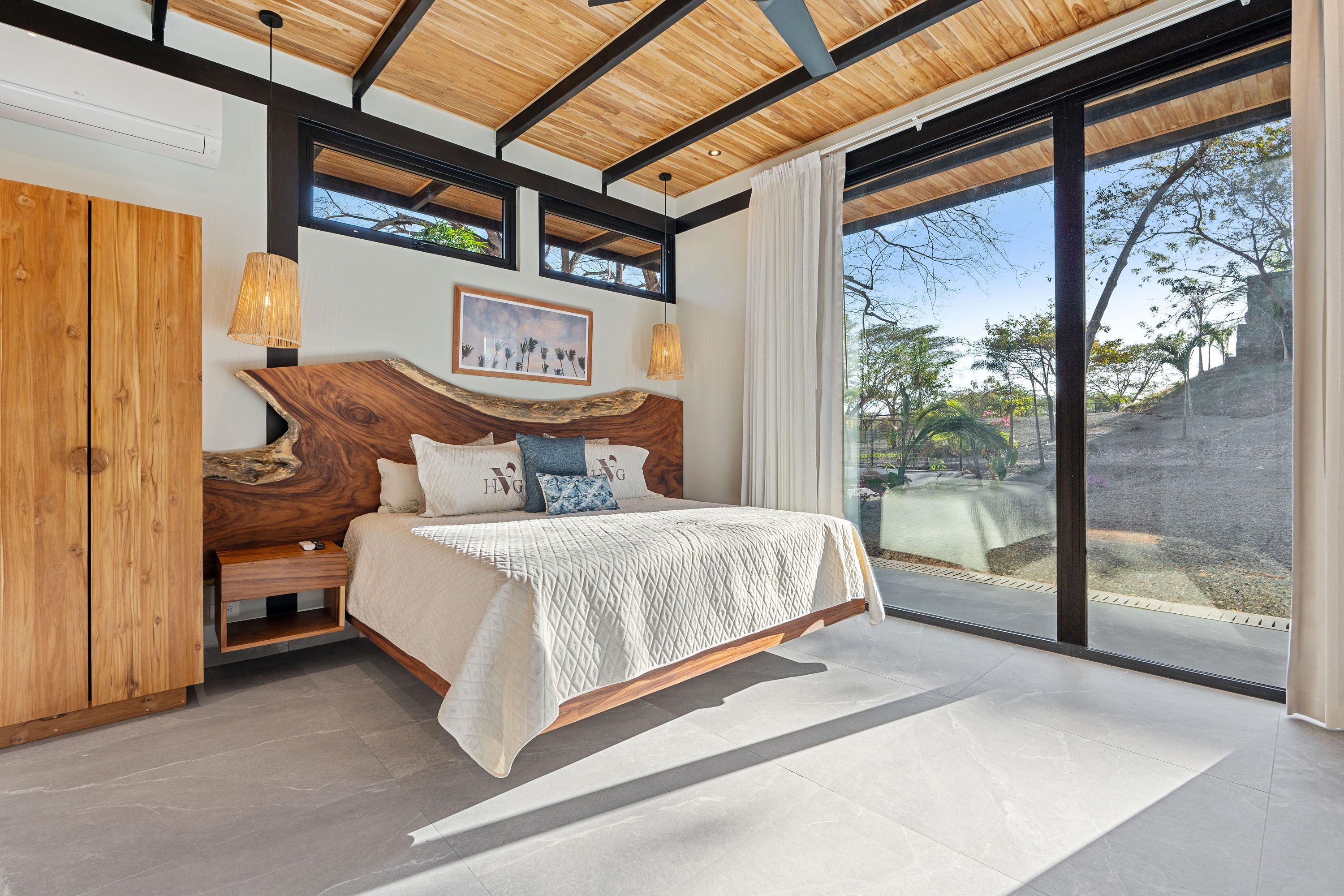 Ein modernes Schlafzimmer mit einem Holz-Kopfteil, einem großen Fenster mit Blick ins Freie und einem Nachttisch mit Lampe.