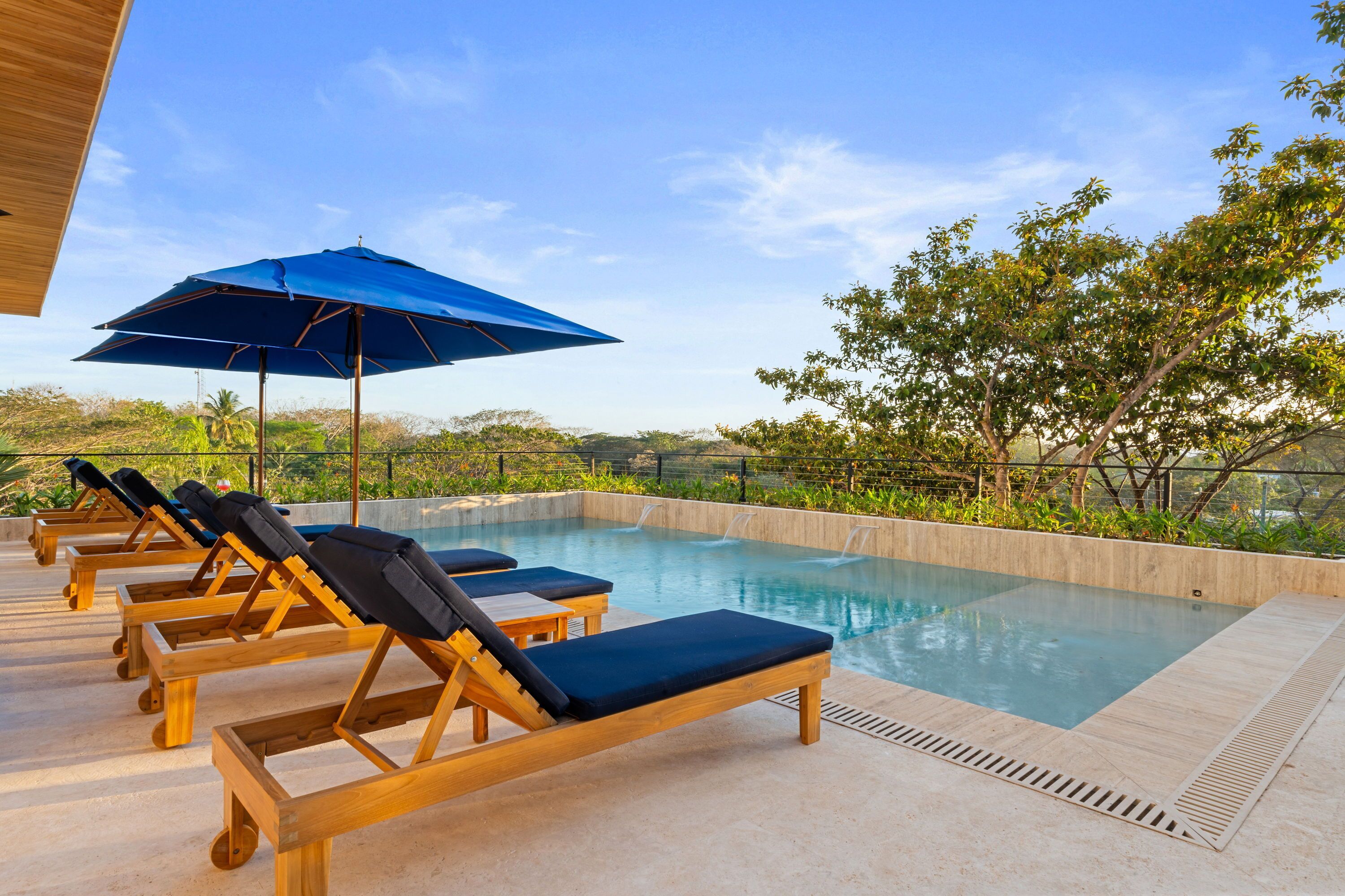 A poolside area with wooden lounge chairs and a blue umbrella, surrounded by greenery and a clear sky.