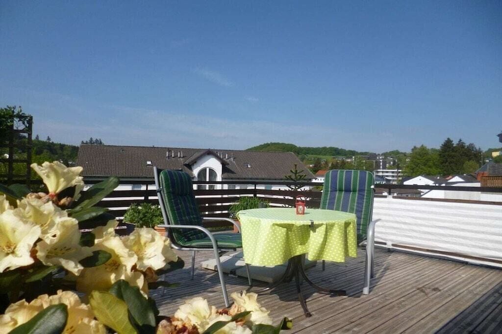 A rooftop terrace with a table and chairs, surrounded by greenery and a view of a building.
