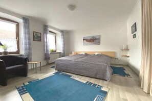 Ein Schlafzimmer mit Bett, Stuhl, kleinem Tisch, einem Fenster mit Vorhängen, einem Bild an der Wand und einem Nachttisch mit Lampe.