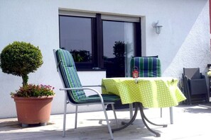 Ein Sitzbereich im Freien mit zwei Stühlen, einem Tisch mit Punkte-Tischdecke, einer Topfpflanze und einem Fenster.