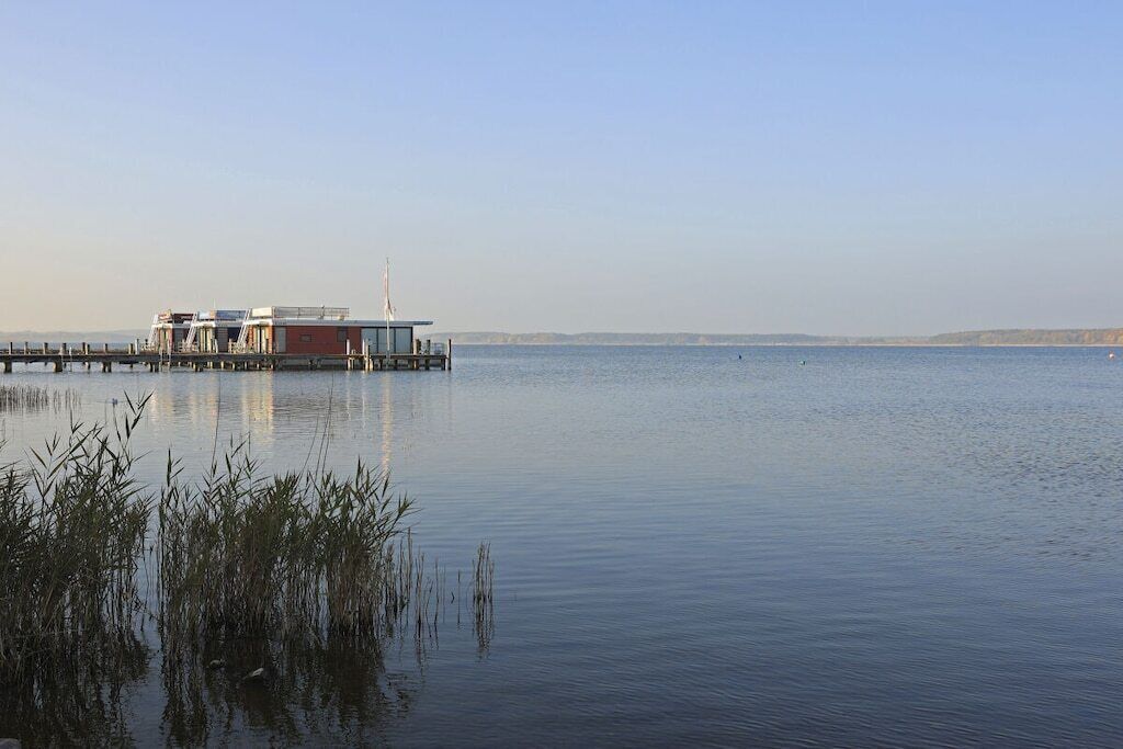 A calm waterfront with a dock extending into the water, a small building with a flag, and reeds in the foreground.