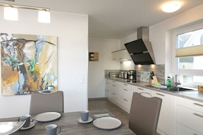 A modern kitchen with white cabinets, a dining area with a table and chairs, and a large abstract painting on the wall. - Villa Paradiso on the lake (Göhren - Lebbin)