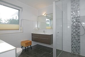 Une salle de bain moderne avec des toilettes blanches, un tabouret en bois, un meuble-lavabo avec un miroir et un espace douche orné de carreaux de mosaïque.
