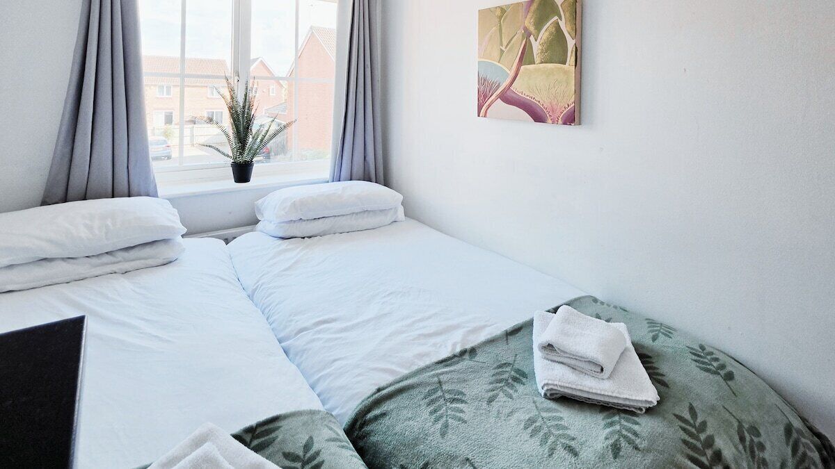 A hotel room with two beds, a window with curtains, a small plant, and a painting on the wall.