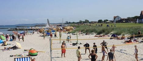 A beach with a volleyball net, beachgoers, and a sailboat in the background.