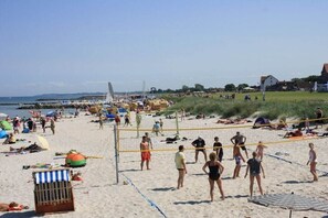 A beach with a volleyball net, beachgoers, and a sailboat in the background. - Schönberger-Strand -Boldt (Schönbergerstrand)