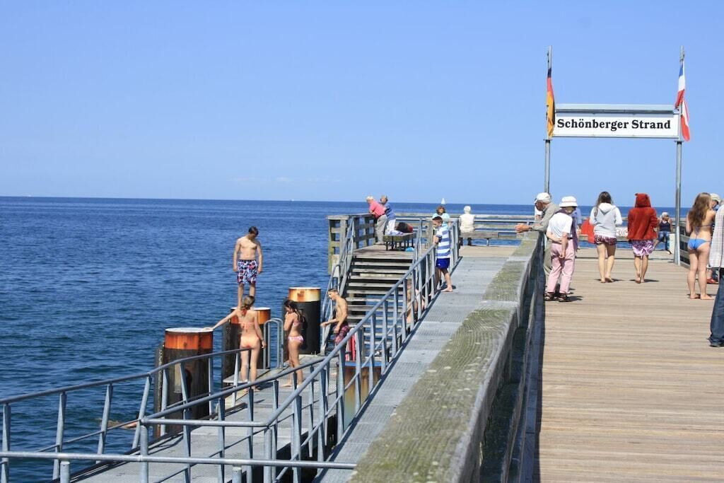 Eine Promenade mit Menschen, die laufen und stehen, ein Schild mit der Aufschrift 'Schönberger Strand' und ein klarer, blauer Himmel.