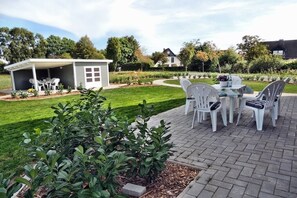 Un espace patio avec une table et des chaises, entouré d’une pelouse bien entretenue et d’un petit abri de jardin.
