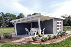 Un espace extérieur aménagé, abrité et doté de tables et de chaises, entouré d’un jardin avec des arbustes et un sentier.