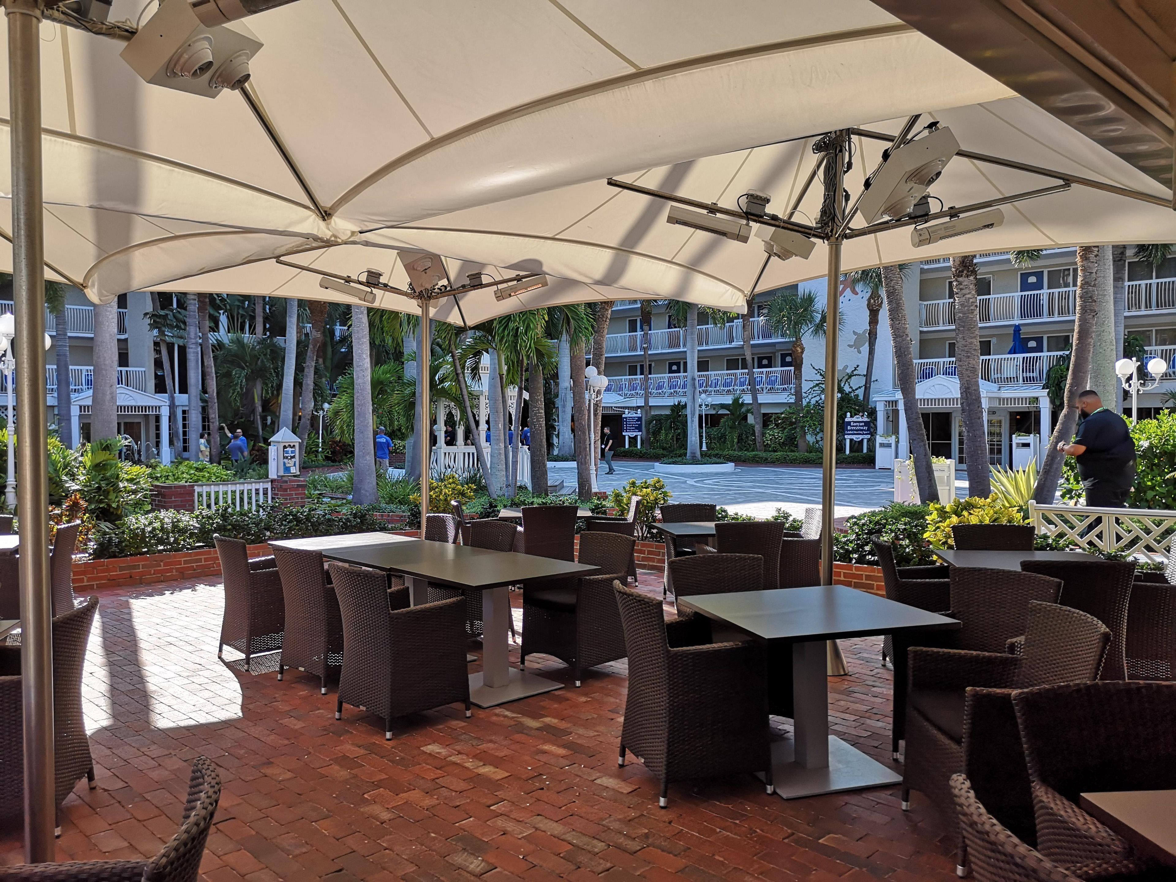 An outdoor patio area with tables and chairs, a brick floor, and a view of a hotel building in the background.