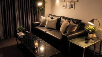 A modern living room with a leather sofa, a glass coffee table, a side table with a lamp, and a large window with curtains.