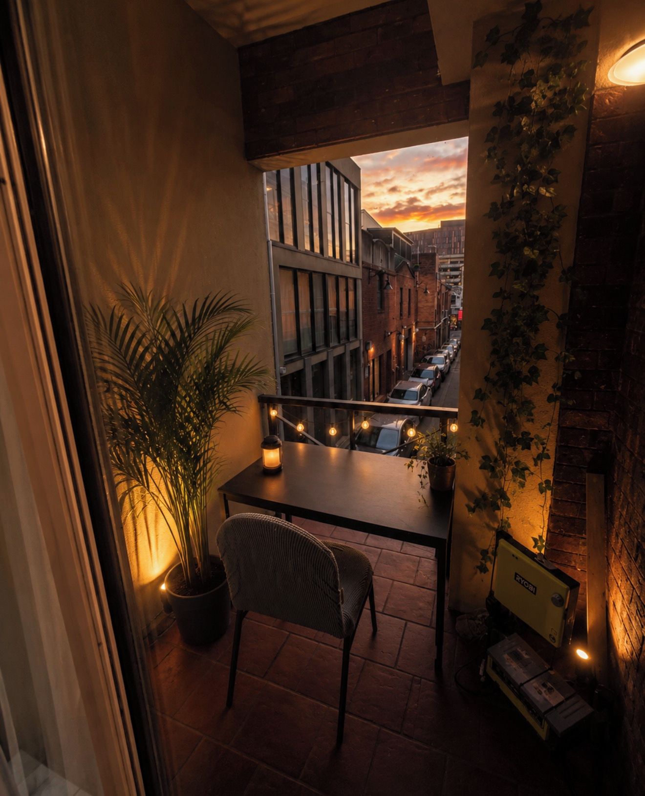 A balcony with a view of a city street at sunset, a table with a chair, and a potted plant.
