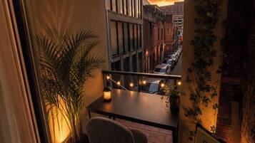 A balcony with a view of a city street at sunset, a table with a chair, and a potted plant.