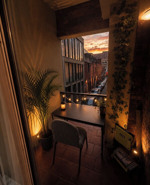 Un balcón con vista a una calle urbana al atardecer, una mesa con una silla y una planta en maceta.