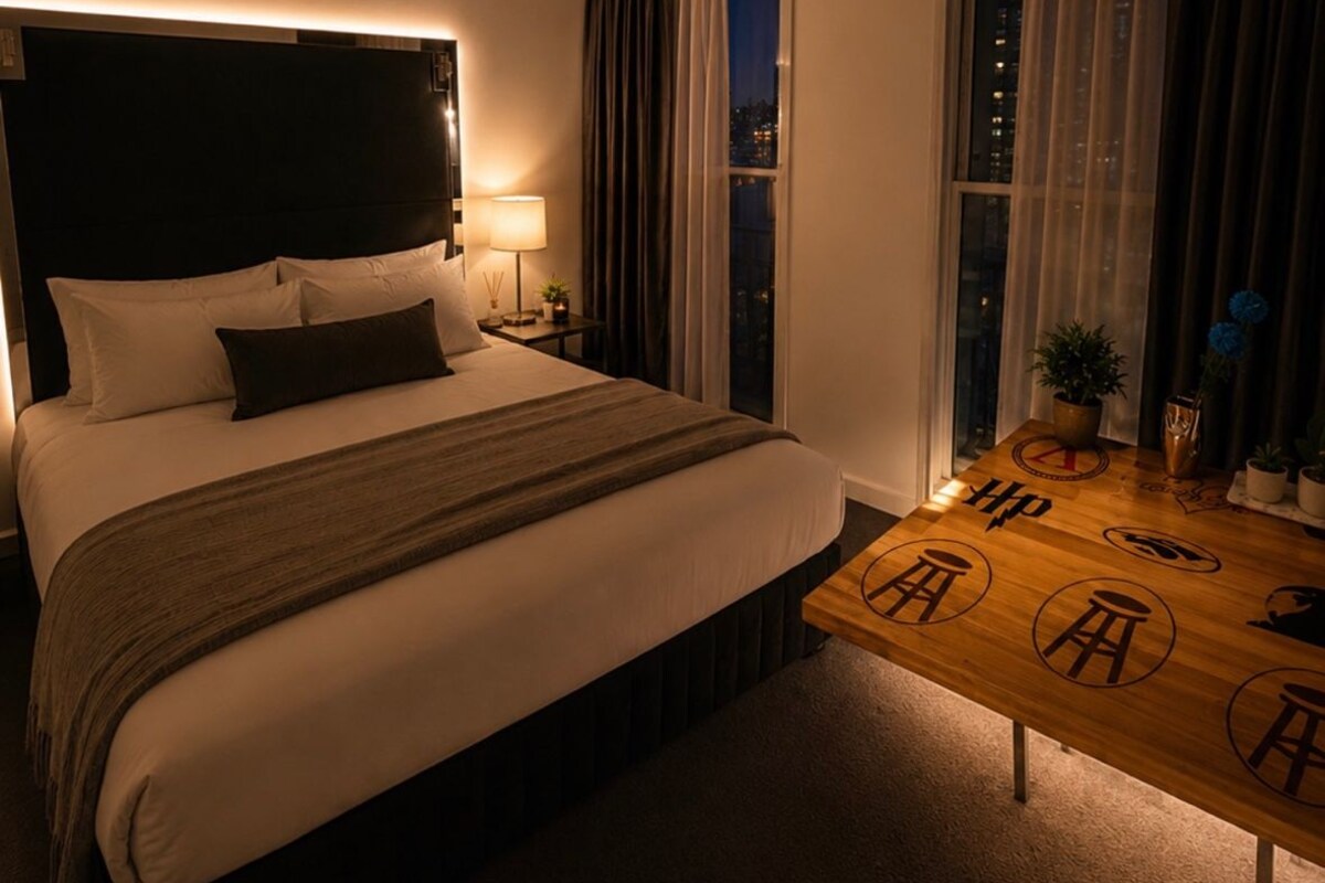 A hotel room with a bed, bedside table, lamp, and a view of the city at night.