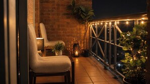 Un balcon avec un mur en briques, deux chaises, une table, des plantes en pot et des guirlandes lumineuses.