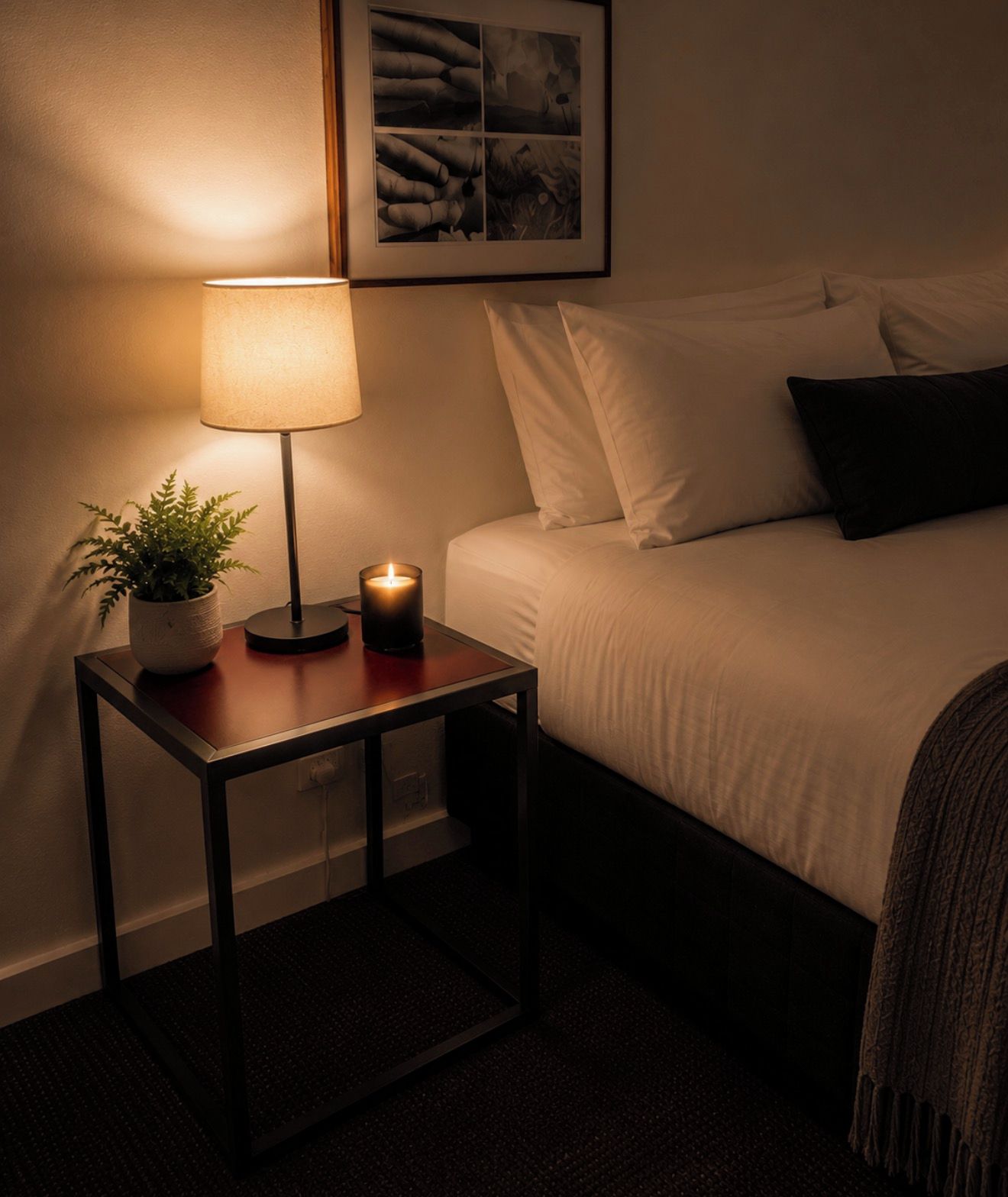 A neatly made bed with white linens, a bedside table with a lamp and a candle, and a framed picture on the wall.