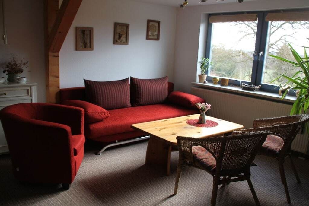 A cozy room with a red sofa, a wooden table, wicker chairs, and a window with plants.