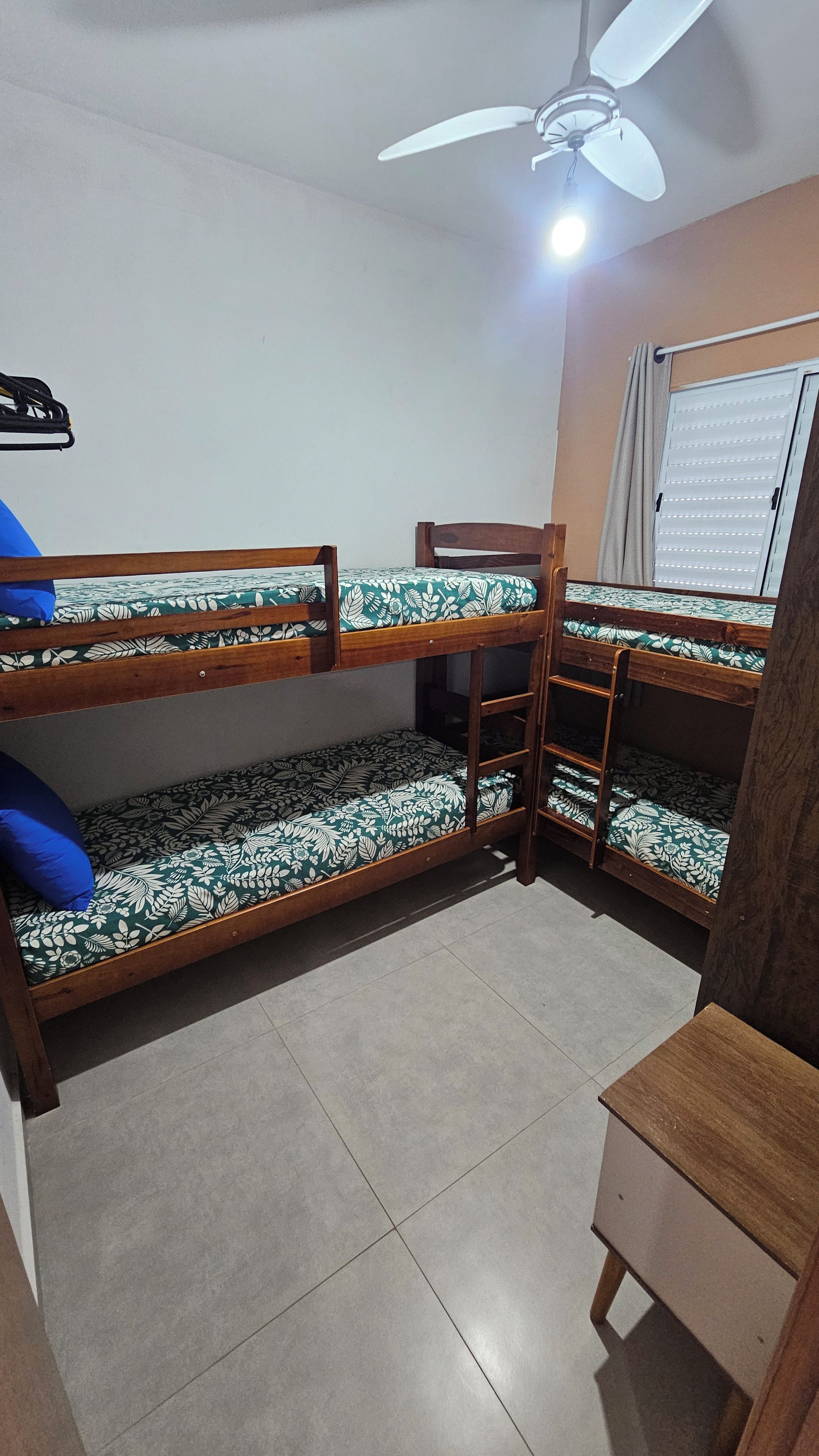 Quarto com beliche, ventoinha no teto, janela com persianas e uma pequena mesa de madeira.