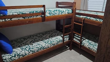 Quarto com beliche, ventoinha no teto, janela com persianas e uma pequena mesa de madeira.