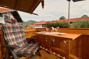 Eine Holzterrasse mit einem für zwei Personen gedeckten Tisch, einem karierten Stuhl und Blick auf Häuser und Grünflächen im Hintergrund.