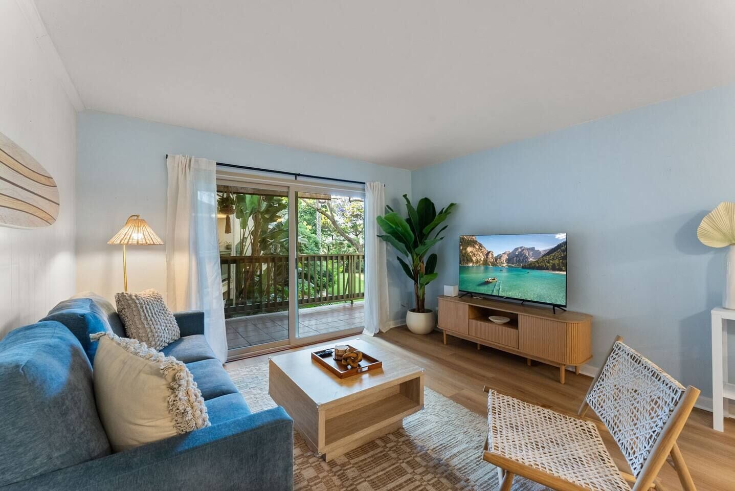 Sala de estar con sofá, mesa de centro, televisor y vista a un paisaje tropical a través de una puerta corrediza.
