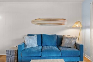 Ein blaues Sofa mit gemusterten Kissen, ein osmanischer Hocker mit geometrischem Muster und ein an der Wand befestigtes abstraktes Kunstwerk.