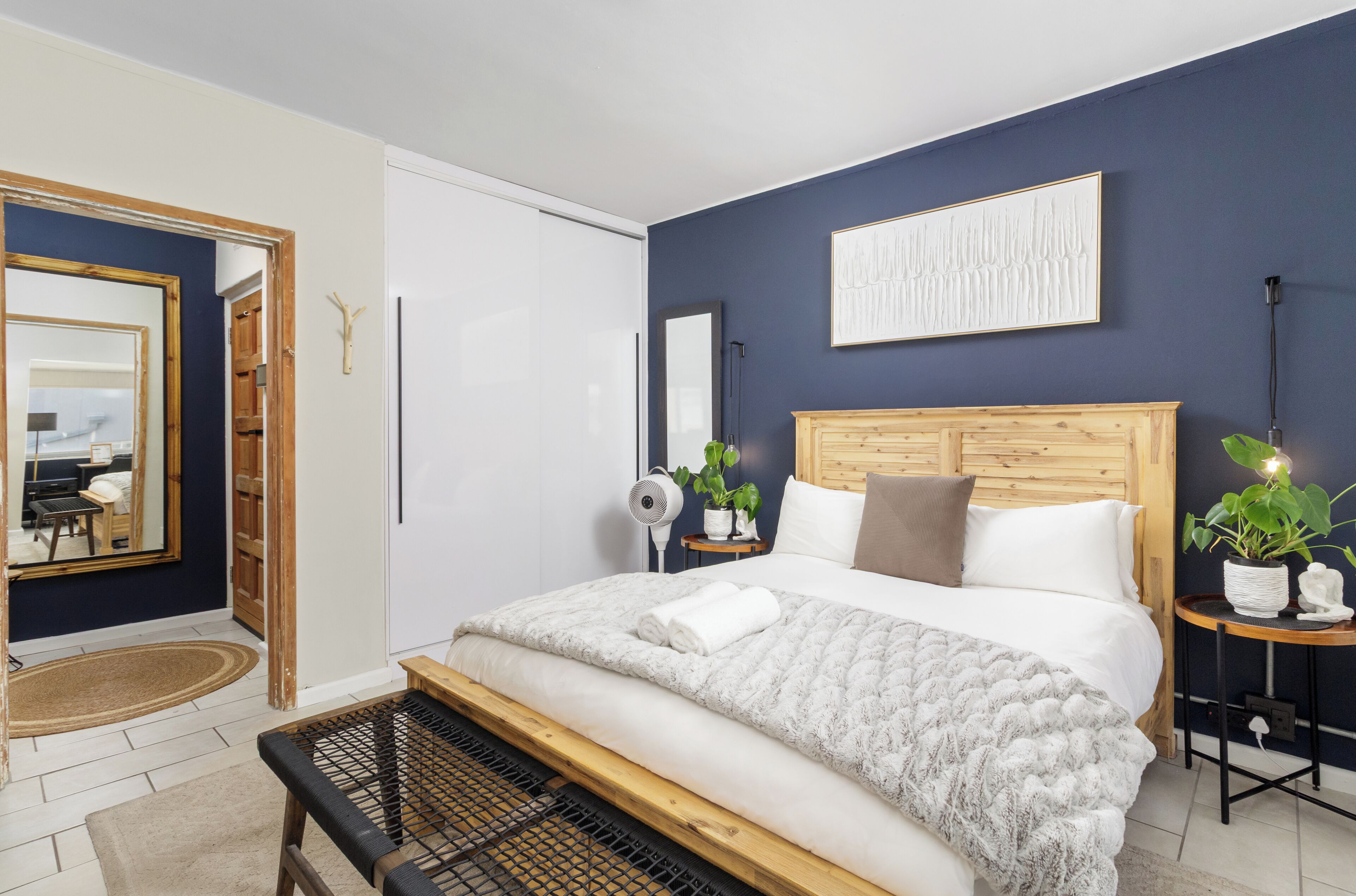 A bedroom with a bed, bedside tables, a mirror, a wall-mounted artwork, and a potted plant.