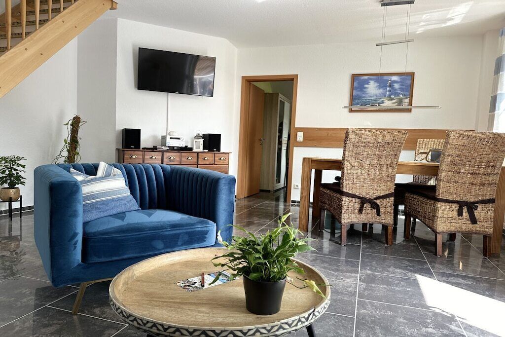 Un salon avec un canapé bleu, une table basse ronde ornée d’une plante, un téléviseur fixé au mur et un coin repas avec des chaises en osier.
