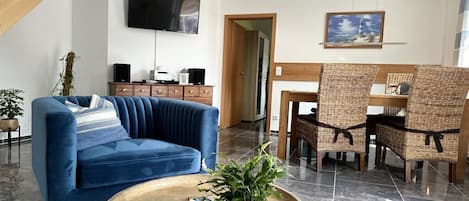 A living room with a blue sofa, a round coffee table with a plant, a TV mounted on the wall, and a dining area with wicker chairs.