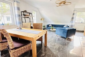 A modern living room with a dining area, featuring a wooden table, wicker chairs, a blue sofa, and a hanging light fixture. - Holiday apartment with 1 bedroom (Ostseebad Prerow)