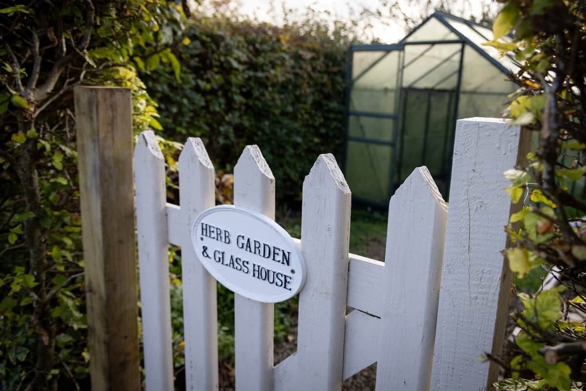 A white picket fence with a sign that reads 'Herb Garden & Glass House.'