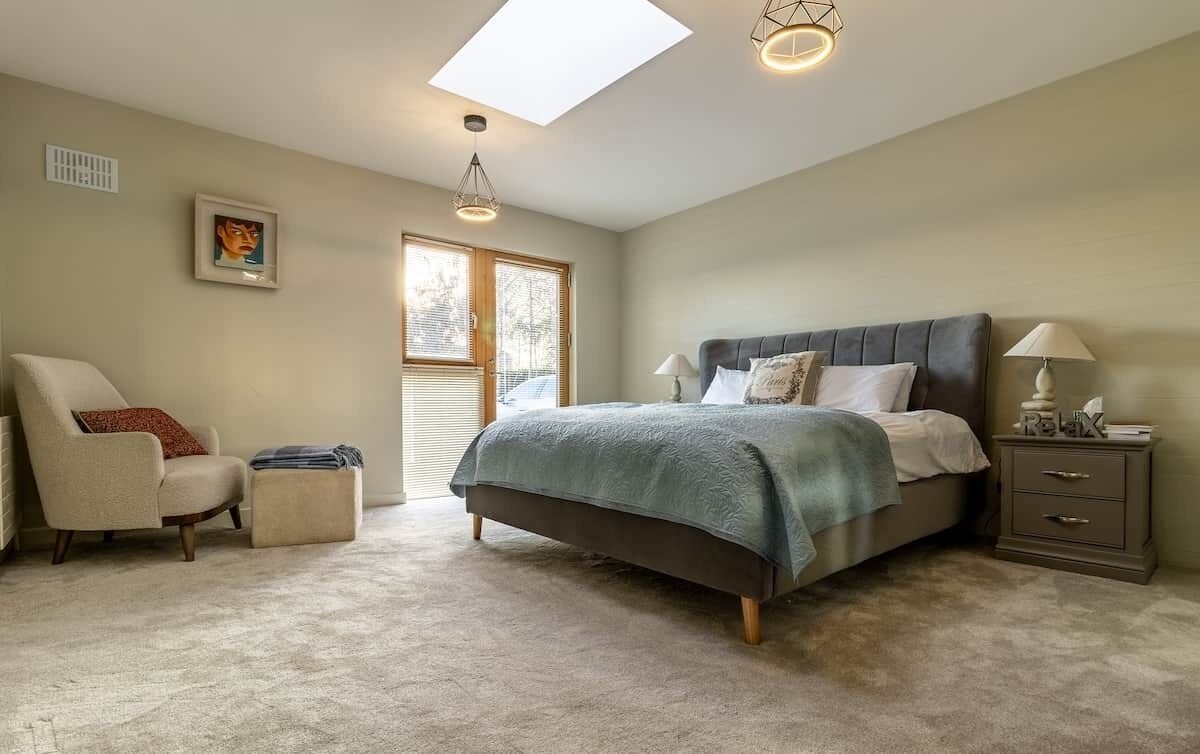Une chambre à coucher comprenant un lit, une chaise, une table de chevet, une lampe, un tableau au mur et un puits de lumière.