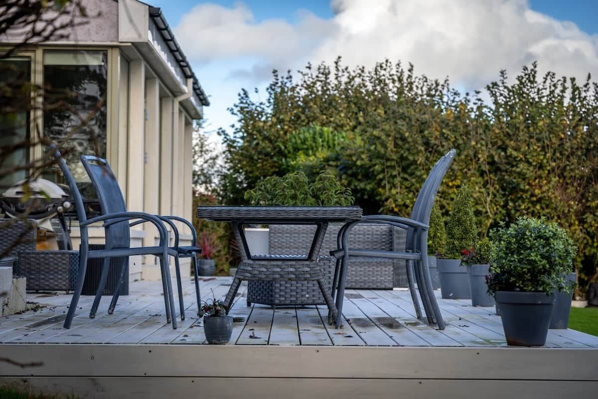 An outdoor patio with a table and chairs, surrounded by potted plants and a hedge.