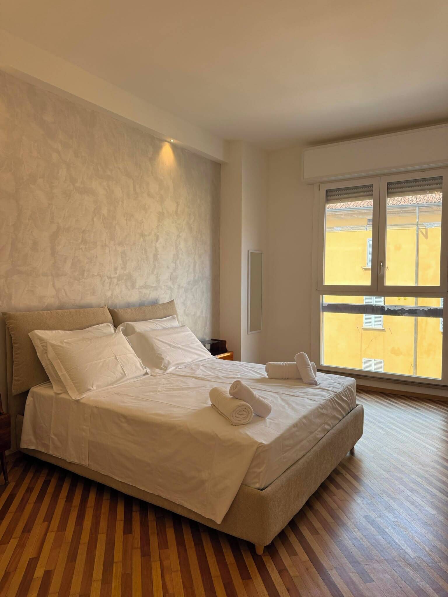 A hotel room with a large bed, wooden flooring, a large window, and a textured wall.